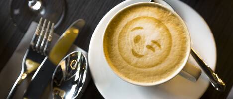 Una taza de café con arte latte sobre un plato pequeño blanco, acompañada de una cuchara y un tenedor sobre una mesa oscura.