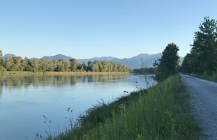 Un río tranquilo con una carretera a la derecha, rodeado de árboles y montañas al fondo.