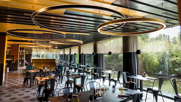 A modern restaurant with circular ceiling lights, large windows with a view of greenery, and a spacious layout with multiple dining tables and chairs. - My Home My Hotel Rosenheim (Rosenheim)