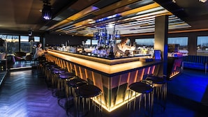 A modern bar area with a long counter, multiple stools, and a bartender. - My Home My Hotel Rosenheim (Rosenheim)