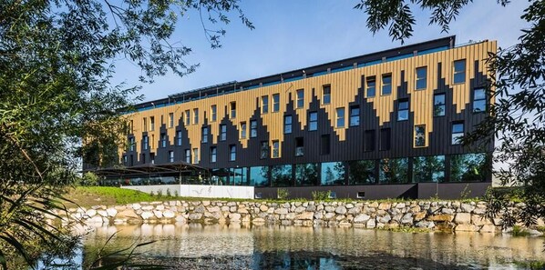 A modern building with a unique facade, a stone wall, and a reflective water body in front. - My Home My Hotel Rosenheim (Rosenheim)