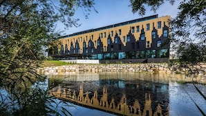 A modern building with a reflective water body in front, surrounded by trees. - My Home My Hotel Rosenheim (Rosenheim)