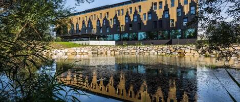 Un bâtiment moderne, avec une étendue d’eau aux reflets changeants à l’avant, entouré d’arbres.