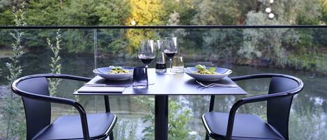 A table set with plates, glasses, and a bottle, overlooking a body of water and greenery.