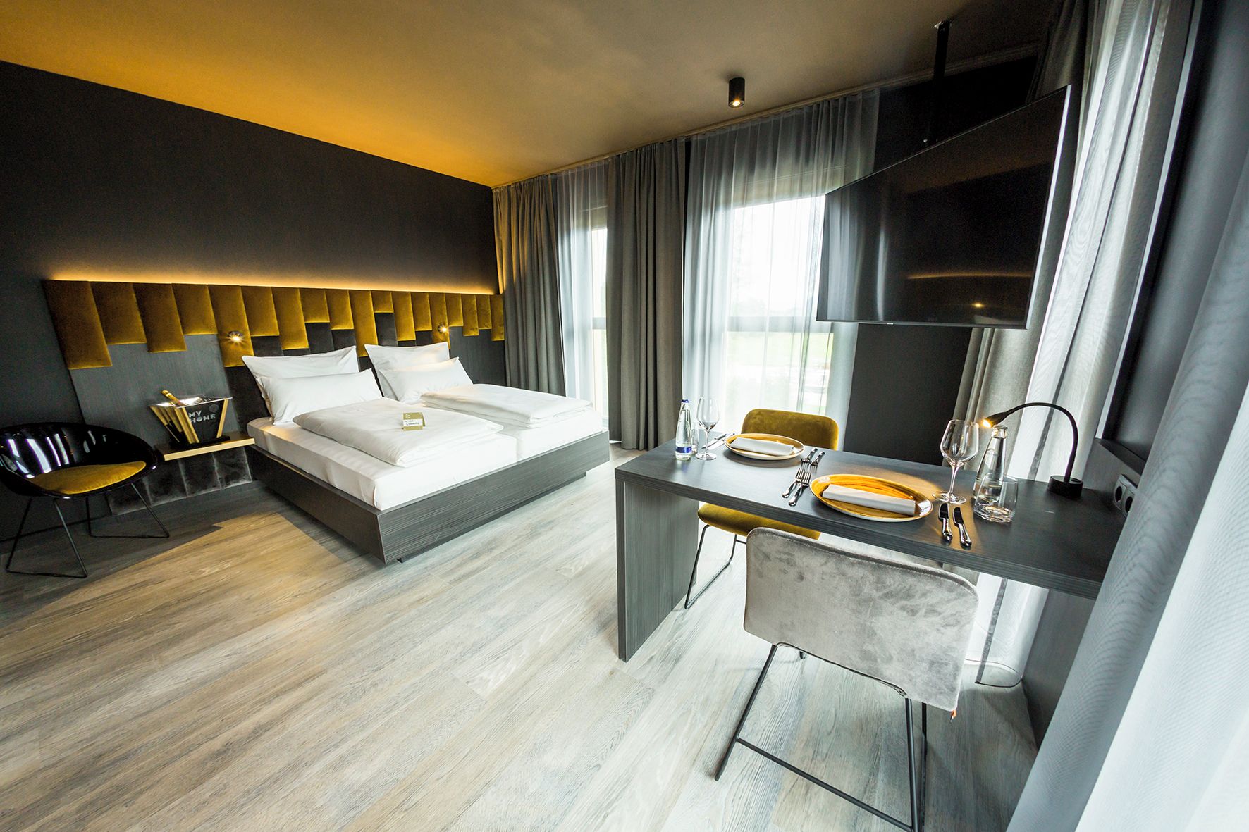 Habitación de hotel moderna con cama, escritorio con silla, minibar y un amplio ventanal con cortinas.