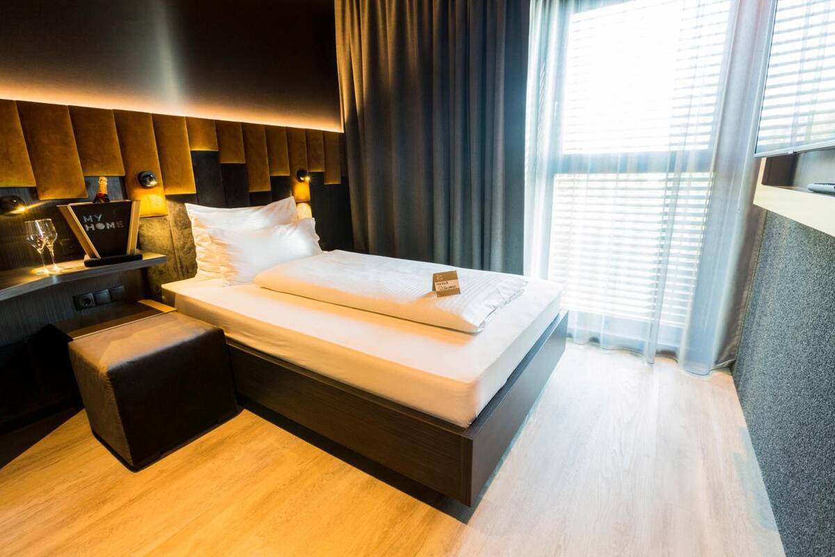Ein modernes Hotelzimmer mit einem großen Bett, einem Nachttisch, einem kleinen Hocker und einem Beistelltisch mit Flasche und Gläsern.