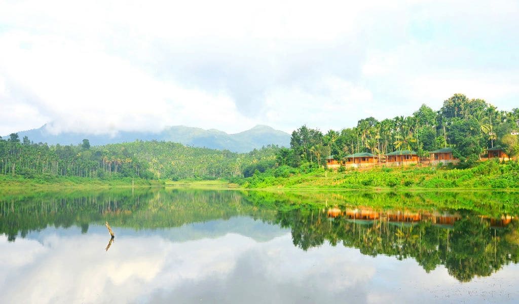 A serene lakeside view with a row of wooden cabins, lush greenery, and a clear reflection on the water.