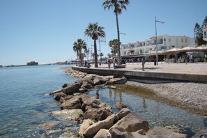 A coastal promenade with a rocky shoreline, palm trees, and buildings with outdoor seating. - Phaedrus Living: Seaside Luxury Flat Lighthouse 68 (Paphos)