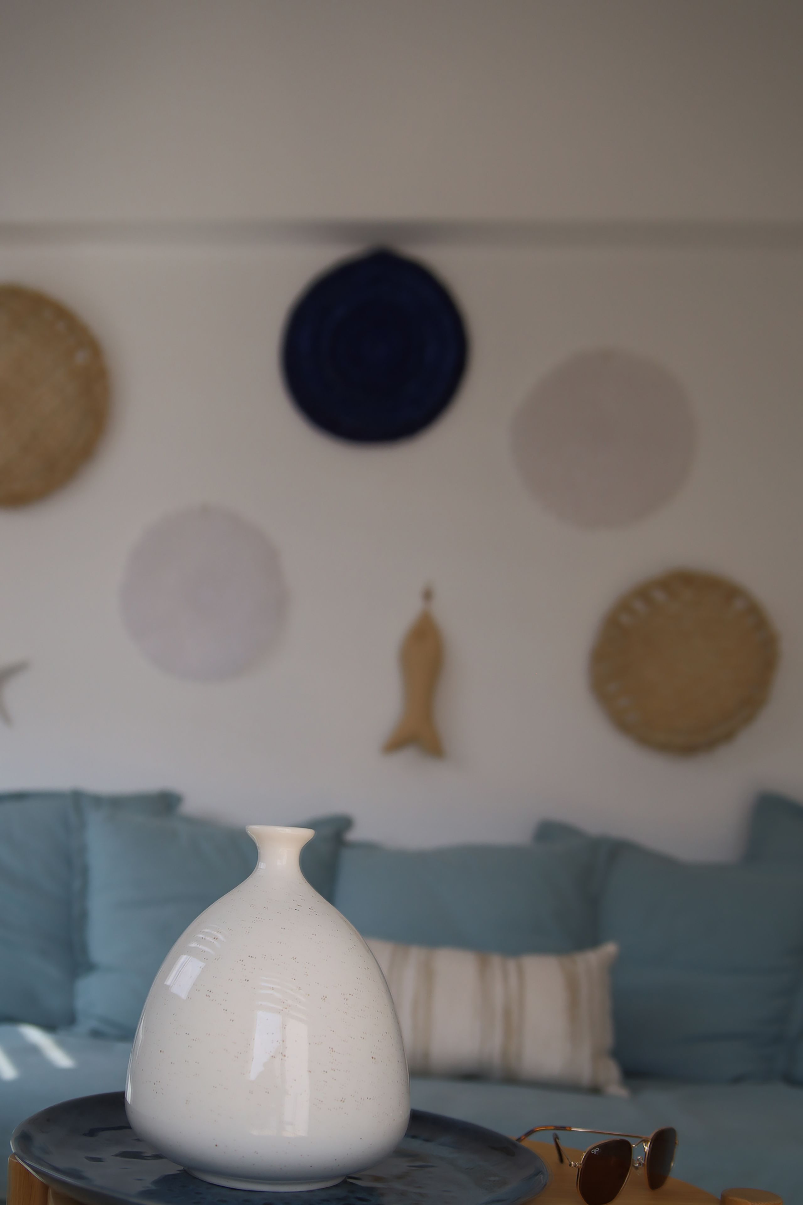 Un jarrón blanco sobre una mesa redonda, un par de gafas de sol y un sofá azul con cojines estampados en una habitación con adornos circulares en la pared.