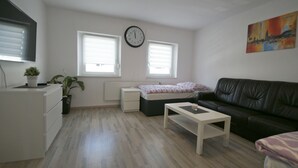 A compact living space with a bed, a sofa, a coffee table, a small desk, and a radiator. There are two windows with blinds, a wall clock, and a painting. - Ferienwohnung  Bergisch Gladbach GL5 (Bergisch Gladbach)
