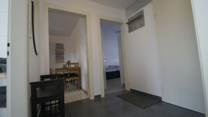 A hallway with a bed in the background, a desk with chairs, and a door with a vent. - Ferienwohnung  Bergisch Gladbach GL5 (Bergisch Gladbach)