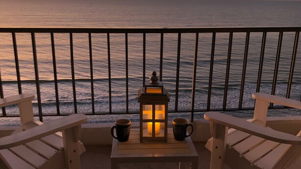 Een balkon met twee Adirondack-stoelen, een kleine tafel en twee mokken. Een lantaarn staat op de tafel. In de verte is de oceaan en een zonsondergang te zien.