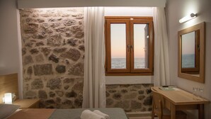 Ein Hotelzimmer mit Steingemäuer, einem hölzernen Bett, einem Schreibtisch und einem Fenster mit Meerblick.