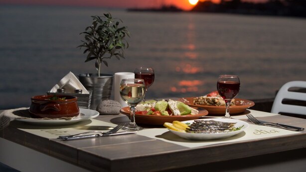 Ein Tisch, gedeckt für ein Abendessen, mit Blick auf einen Sonnenuntergang, inklusive einer Pflanze, Weingläsern und Tellern mit Speisen.