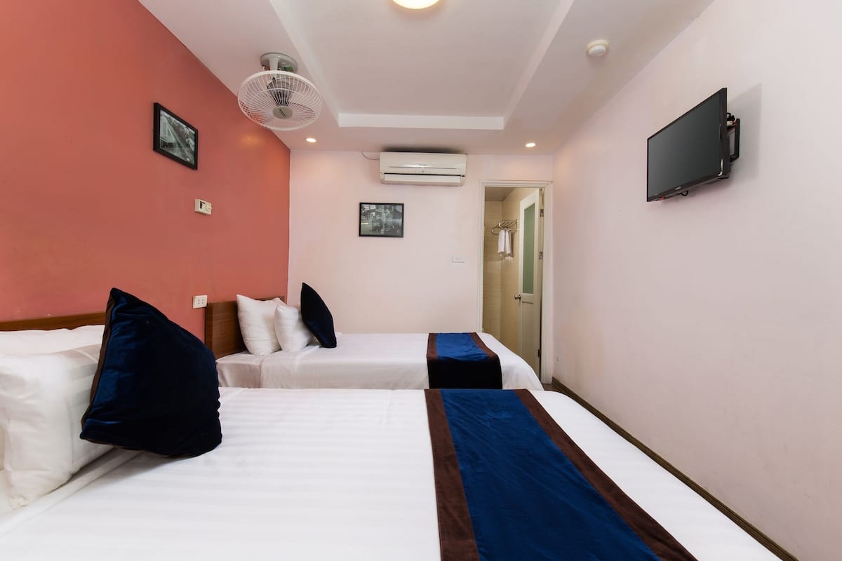 Habitación de hotel con dos camas, ventilador de techo, unidad de aire acondicionado, televisor de pantalla plana y puerta que da al baño.