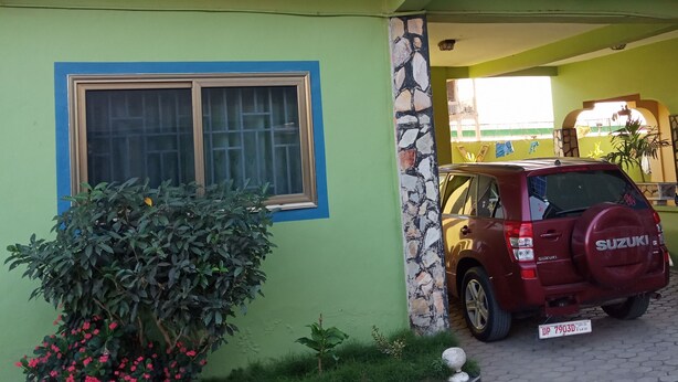 A two-story building with a green facade, a large window with a blue frame, and a parked red Suzuki SUV.