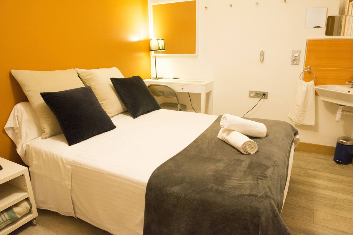 A hotel room with a bed, a desk, a chair, a lamp, a sink, and a mirror.