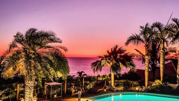 Zona de piscina con vista al atardecer, rodeada de palmeras y una terraza de madera.