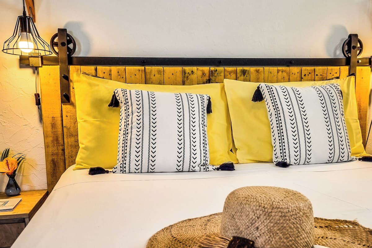 Un lit avec une tête de lit en bois, des oreillers jaunes et un chapeau de paille posé sur le lit.