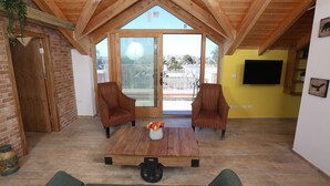 Un vaste espace de vie doté d’un haut plafond et de poutres en bois, une grande porte-fenêtre coulissante donnant sur un patio extérieur, deux fauteuils à rayures, une table basse agrémentée d’un vase de fleurs et un téléviseur à écran plat fixé au mur.
