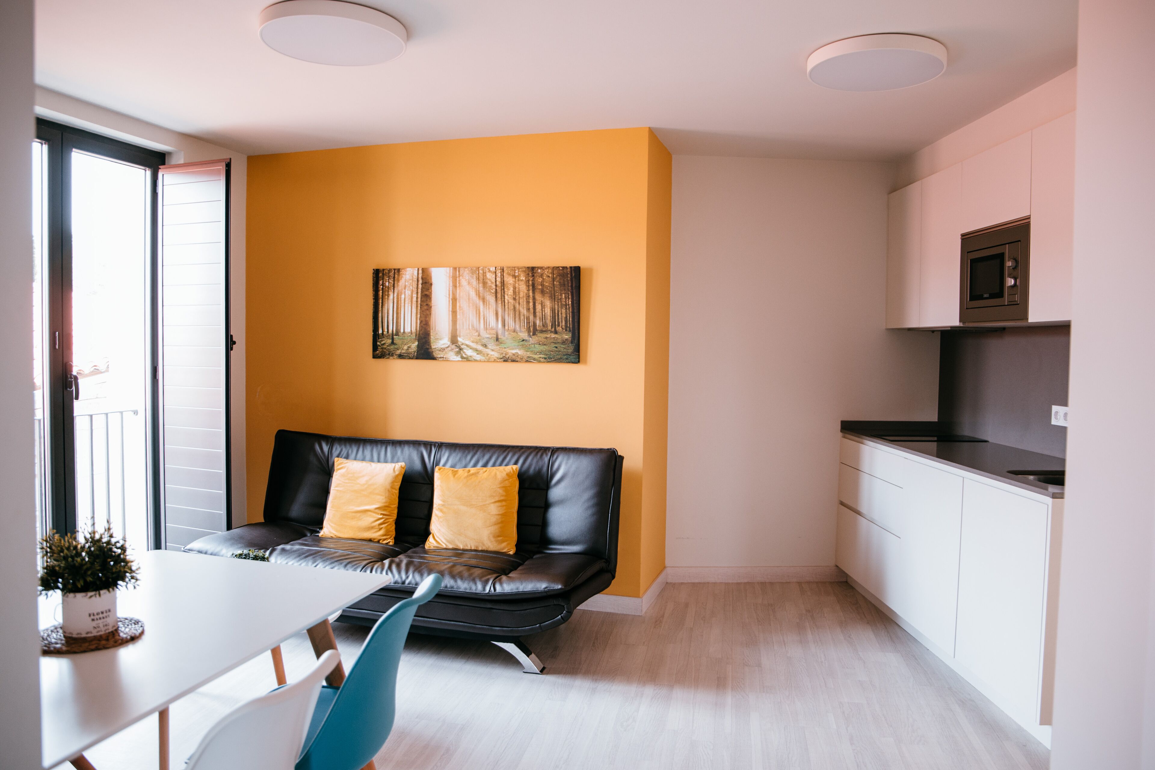 A modern kitchen with white cabinets and a microwave, a black leather sofa with yellow cushions, a white dining table with a blue chair, and a painting of a forest on the wall.