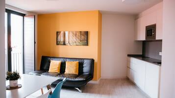 A modern kitchen with white cabinets and a microwave, a black leather sofa with yellow cushions, a white dining table with a blue chair, and a painting of a forest on the wall.