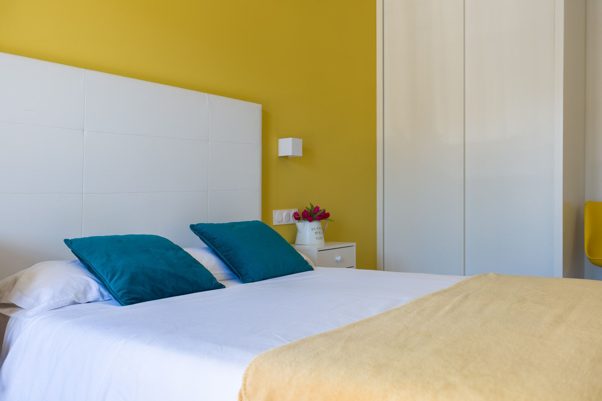 A bed with white bedding and teal pillows, a white nightstand with a vase of flowers, and a yellow wall.