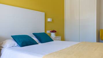 A bed with white bedding and teal pillows, a white nightstand with a vase of flowers, and a yellow wall.