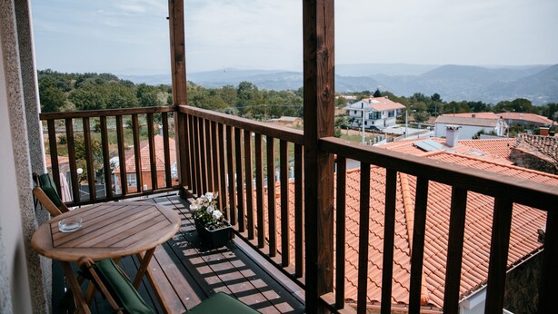 A wooden balcony with a table and chairs, overlooking a cityscape with red-tiled roofs and distant mountains.