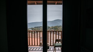 A view from a balcony with a wooden railing, a chair, and a table, overlooking a landscape with hills and trees.