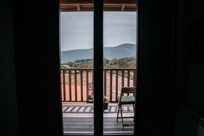 A view from a balcony with a wooden railing, a chair, and a table, overlooking a landscape with hills and trees.