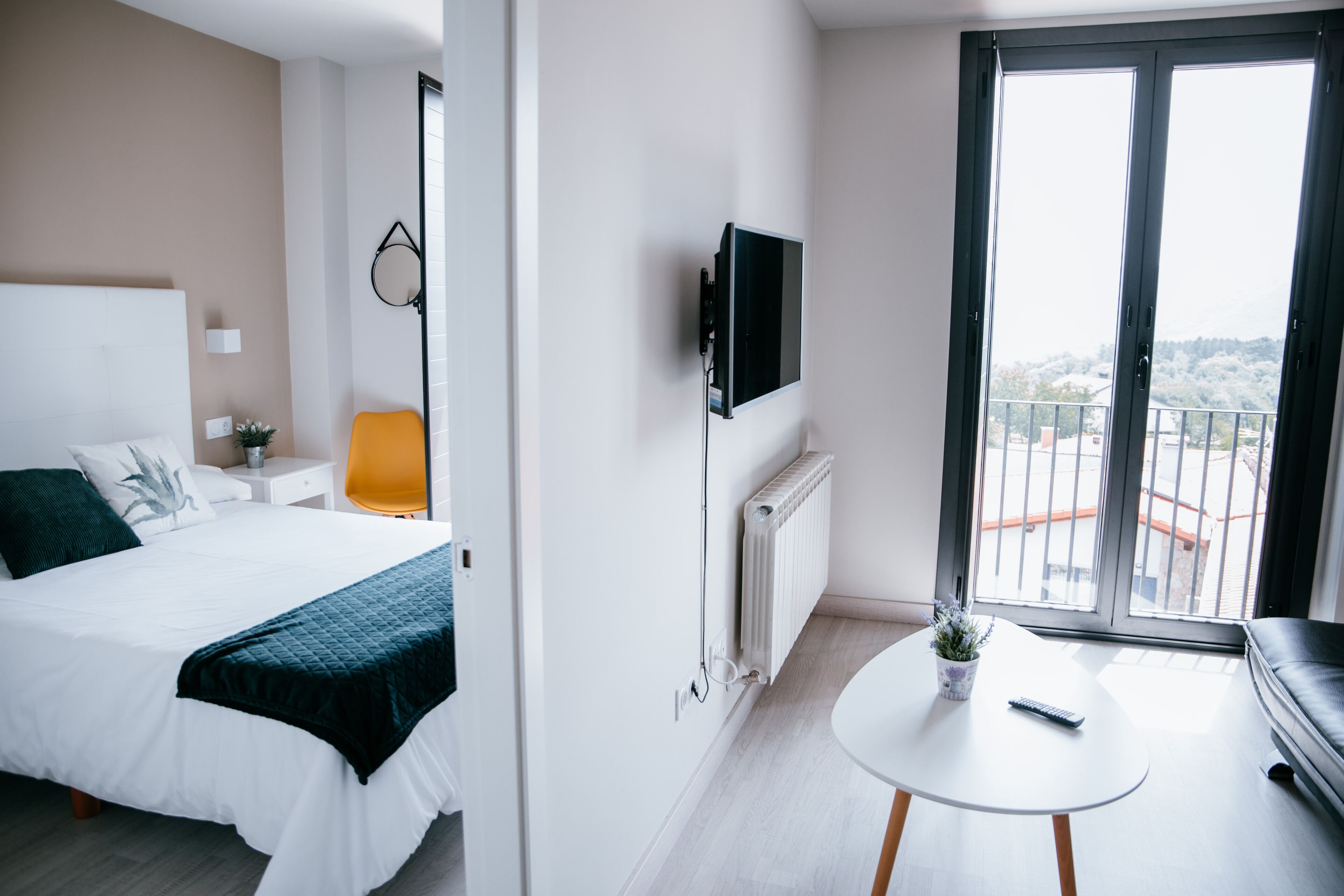 A modern hotel room with a large bed, a TV mounted on the wall, a small table with a remote control, and a balcony with a view.