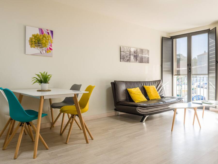 A modern living room with a dining area, a sofa with yellow cushions, and a large window offering a view of the outside.