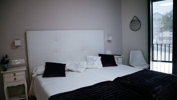 A bedroom with a large bed, a bedside table, a nightstand, a chair, and a window with a view of the outside.