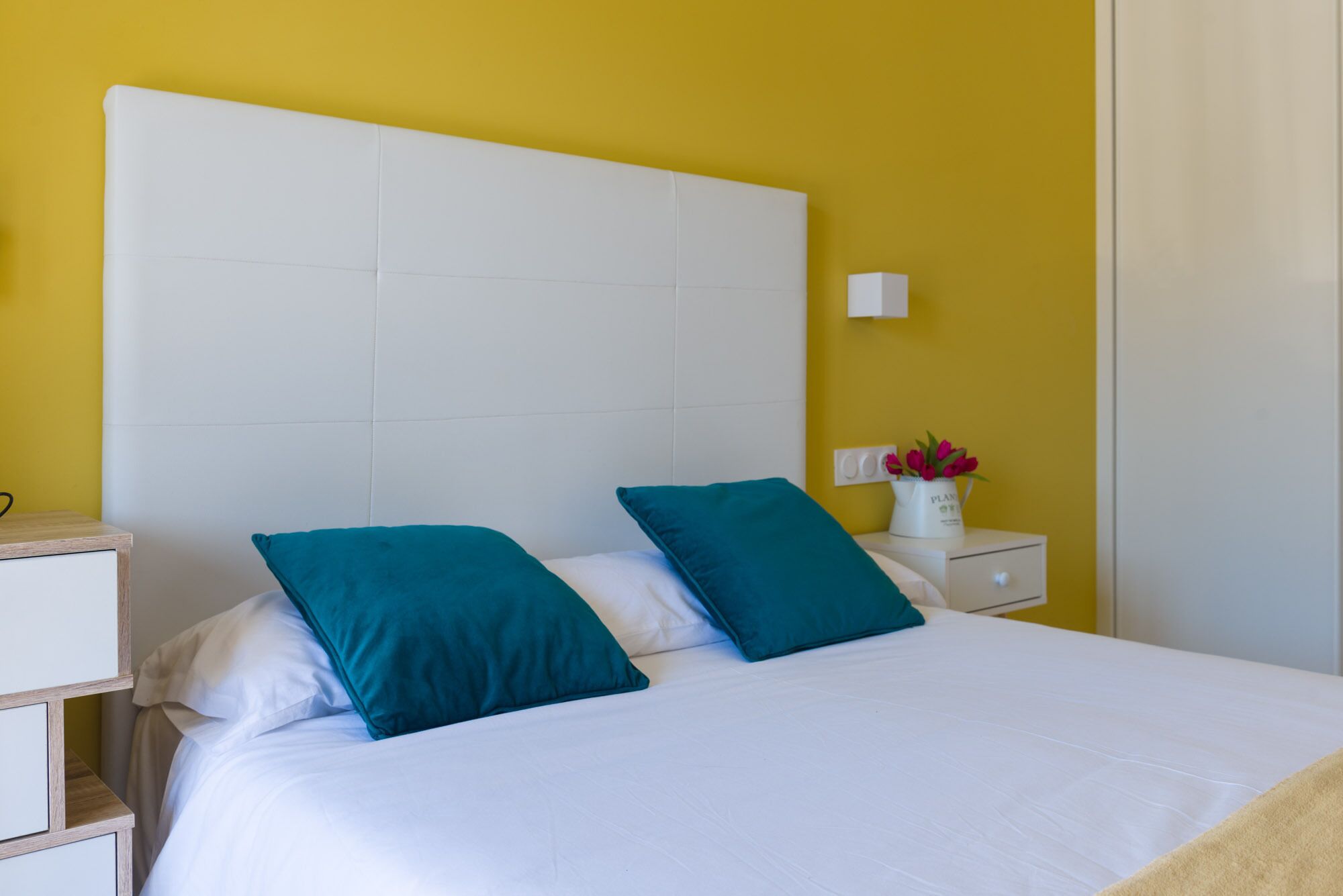 Una cama con ropa blanca y almohadas turquesa, una mesita de noche blanca con un florero de flores y una pared amarilla.