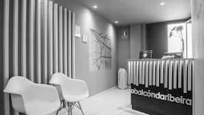 Un área de recepción con diseño moderno, que incluye un mostrador de recepción con un mapa y un cartel, dos sillas blancas y un jarrón decorativo.