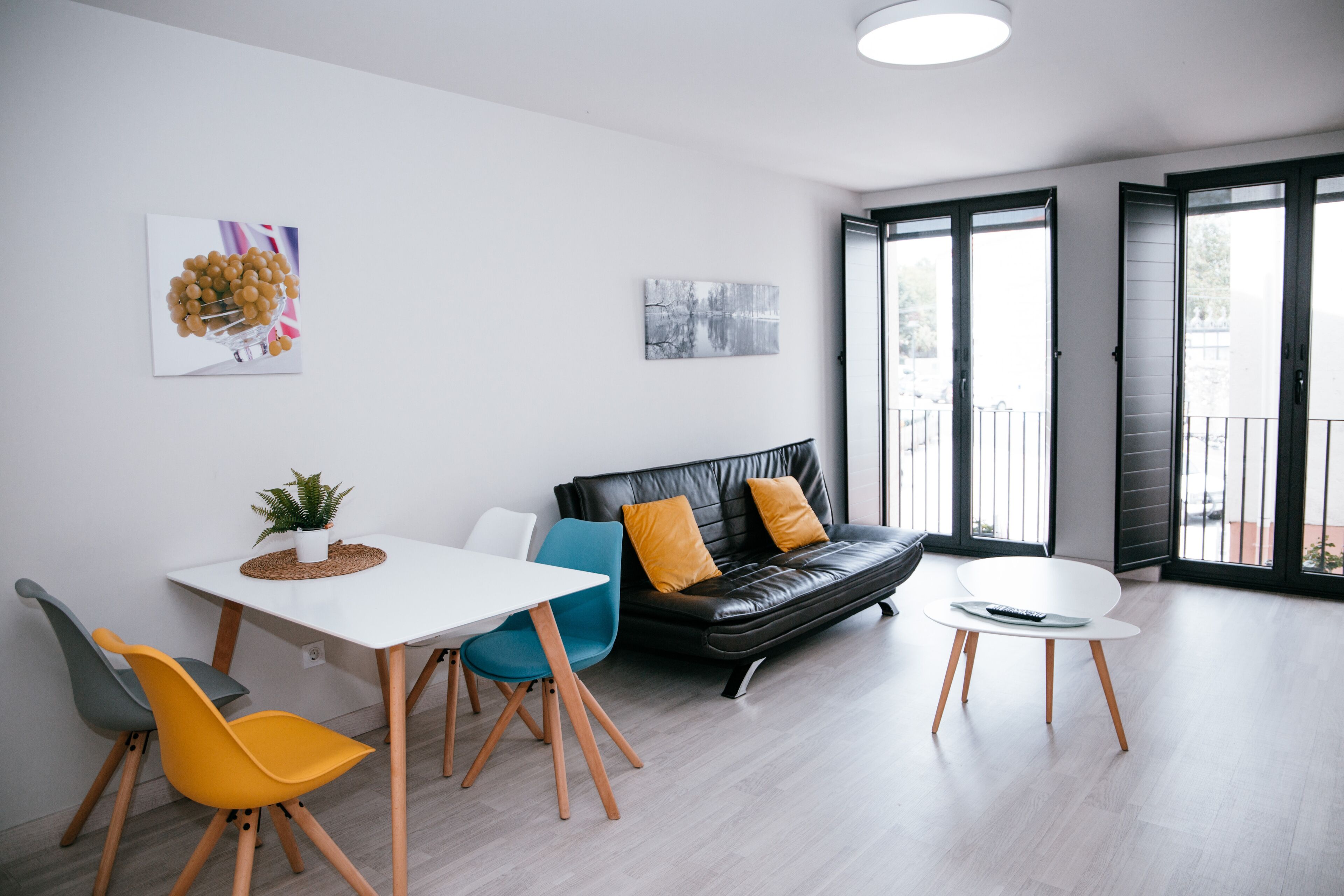 Un salón moderno con un sofá de cuero negro, una mesa blanca y dos sillas. Hay una planta pequeña sobre la mesa y obras de arte en las paredes.