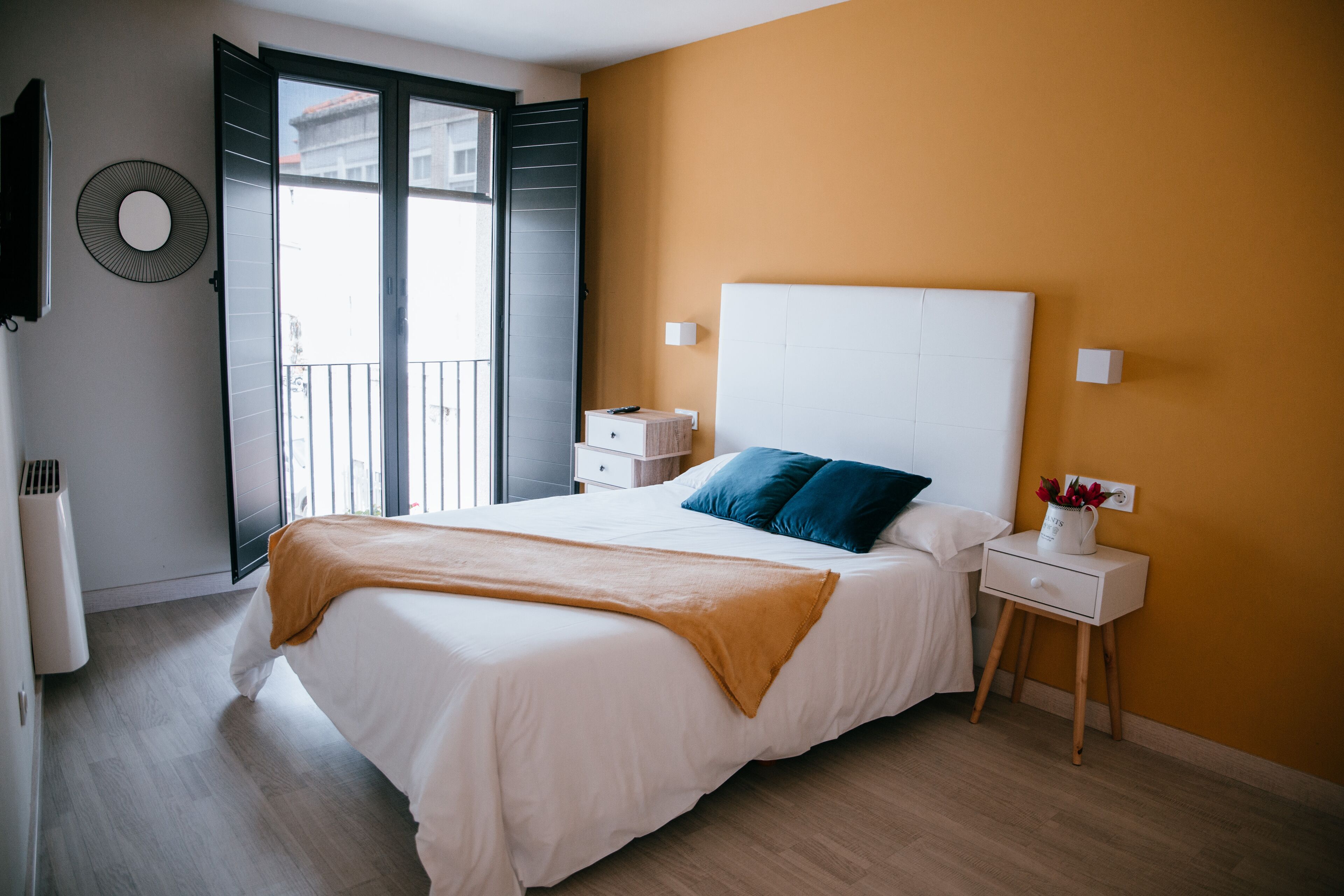 A bedroom with a bed, a nightstand, a wall-mounted TV, and a window with a view of buildings.