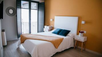 A bedroom with a bed, a nightstand, a wall-mounted TV, and a window with a view of buildings.