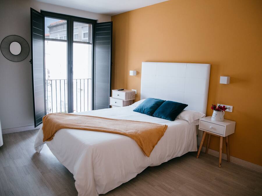 A bedroom with a bed, a nightstand, a wall-mounted TV, and a window with a view of buildings.