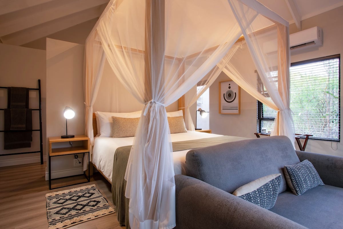 A bedroom with a canopy bed, a grey sofa, a bedside table with a lamp, and a patterned rug.