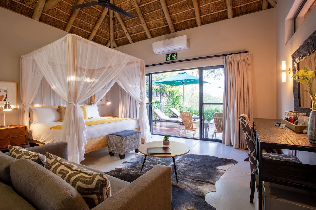 A bedroom with a four-poster bed, a sitting area with a table and chairs, and a view of the outdoors through large windows.