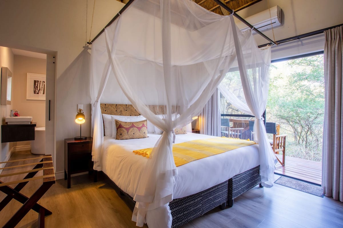 A bedroom with a canopy bed, a bedside table with a lamp, a wooden floor, and a view of the outdoors through a large window.