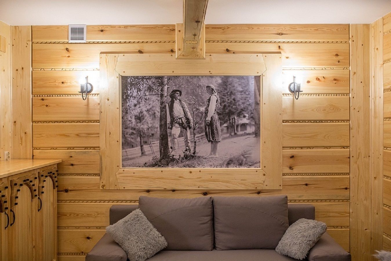 A wooden cabin interior with a framed black and white photograph, a sofa, and two wall-mounted lights.