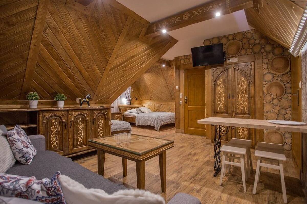 A cozy attic bedroom with a loft bed, a small dining table, and a TV mounted on the wall.