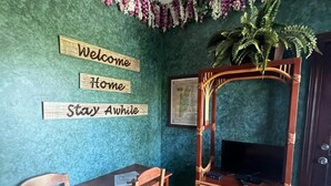 A room with a green textured wall, a wooden TV stand with a flat-screen TV, two wooden chairs, a small table, and a welcome sign. - Belize Budget Suites (San Pedro)