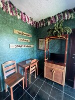 Een kamer met een groene, gestructureerde wand, een houten tv-meubel met een flatscreen televisie, twee houten stoelen, een klein tafeltje en een welkomstbord.