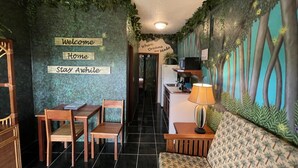 A cozy room with a dining area, a sofa, and a kitchen area. There are wooden chairs and a table in the dining area, and a lamp on a side table. The walls are decorated with greenery and a mural. - Belize Budget Suites (San Pedro)