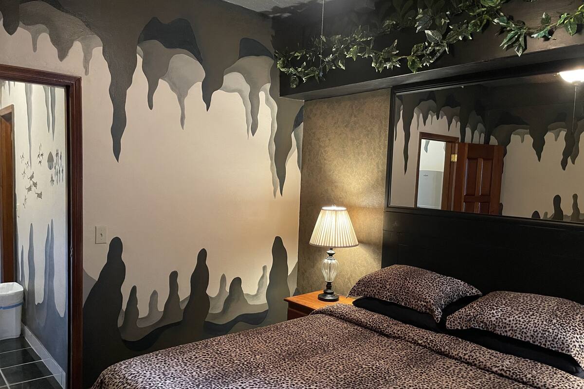 A bedroom with a bed, a nightstand, a lamp, and a wall with a dripping paint design.