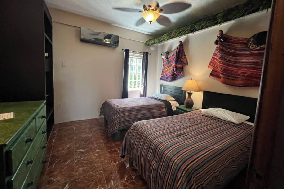 A room with two beds, a ceiling fan, a window with curtains, and a green dresser.